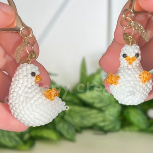 May include: Two handmade white beaded duck earrings with orange beaks and feet. The duck earrings are held up by gold-colored earring hooks. The earrings are in front of a blurred green background.
