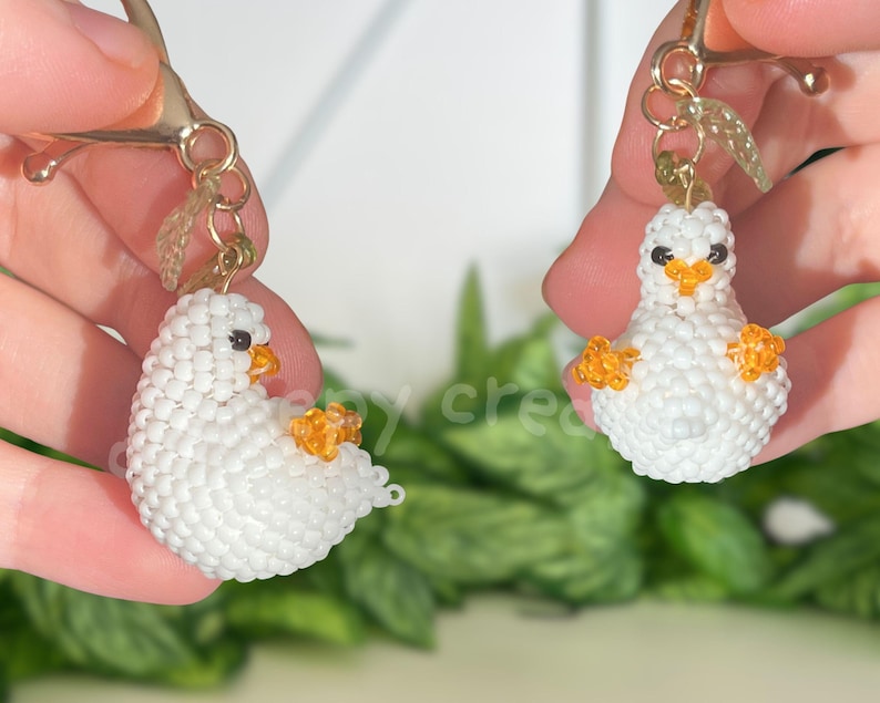 May include: Two handmade white beaded duck earrings with orange beaks and feet. The duck earrings are held up by gold-colored earring hooks. The earrings are in front of a blurred green background.
