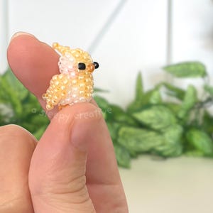 May include: A miniature bird figurine crafted from small, glossy yellow and pale peach beads. The bird features a small crest on its head, two tiny black beads for eyes, and a rounded body shape. The text a sleepy creative is visible.