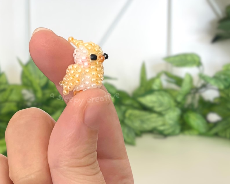 May include: A miniature bird figurine crafted from small, glossy yellow and pale peach beads. The bird features a small crest on its head, two tiny black beads for eyes, and a rounded body shape. The text a sleepy creative is visible.