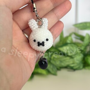May include: A white beaded bunny charm with black eyes and a black bow tie. The bunny is attached to a black cord with a silver-colored clasp. A black, round bead hangs below the bunny. The text "a sleepy creative" is visible.