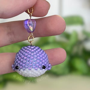 May include: A handmade beaded whale earring in shades of purple and white. The whale is crafted with small beads, featuring a heart-shaped bead and gold-colored metal findings. The earring is held up by a hand, with a blurred green background.
