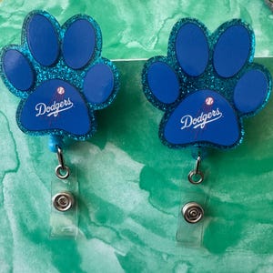 以下が含まれることがあります： グリッター仕上げの青い肉球型バッジリール2個。ドジャースのロゴと野球のグラフィックが描かれています。各バッジリールには、身分証や鍵を安全に取り付けるための金属製スナップボタン付きの透明なプラスチックストラップが付いています。