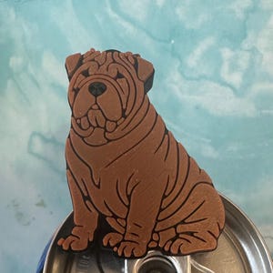 May include: A brown Shar-Pei dog figurine with prominent skin folds and a black nose, eyes, and outline, positioned on top of a silver metal beverage can lid. The dog is depicted in a seated posture, showcasing detailed textured lines.