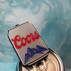 Op de afbeelding: Een zilverkleurige flesopener in de vorm van een blikje met het Coors-logo in rood en blauw. De opener ligt op een Coors-bierblikje, tegen een lichtblauwe en witte achtergrond.