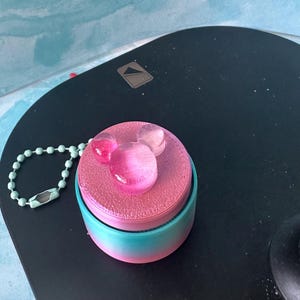 May include: A pink and blue ombre container with a Mickey Mouse design on top. The Mickey Mouse design is made of clear, pink-tinted resin. A light blue beaded chain is attached to the container.