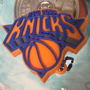 Puede incluir: Llavero con el logotipo naranja y azul de los New York Knicks. El logotipo presenta el nombre del equipo y una pelota de baloncesto. El llavero tiene una cadena de cuentas azules.