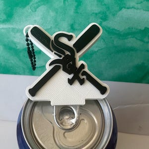 Può includere: Un portachiavi stampato in 3D bianco e nero con un design a mazze da baseball incrociate e il logo dei Chicago White Sox al centro. Il portachiavi è fissato a una catenella a palline nera ed è posizionato sopra la linguetta di una lattina.