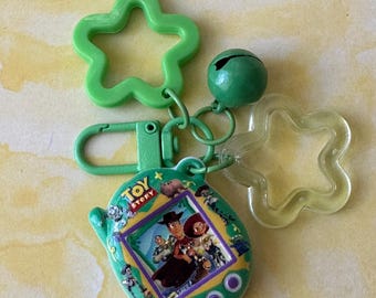 Könnte beinhalten: Ein grüner Kunststoff-Schlüsselanhänger mit einem Toy Story-Digital-Haustier-Spielzeug, dessen Bildschirm Woody und Jessie zeigt. Der Anhänger umfasst einen grünen sternförmigen Anhänger, einen transparenten Stern, eine grüne Glocke und einen Karabiner.