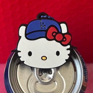 May include: A white Hello Kitty character charm with a blue baseball cap featuring a logo and a red bow, attached to a blue ball chain. The charm is positioned over the silver pull tab of a blue aluminum beverage can against a red background.