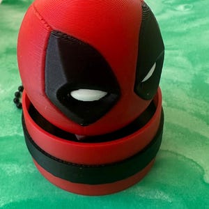 May include: A 3D printed spherical object featuring the red and black mask of the character Deadpool. The top half is a round red head with black eye patches and white eyes, resting inside a matching red and black cylindrical base container.