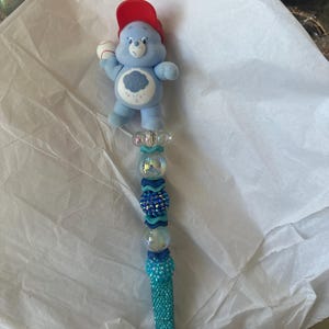 May include: A decorative pen featuring a light blue Care Bear figure wearing a red baseball cap and holding a baseball. The pen's shaft is adorned with blue and clear beads, and the pen tip is covered in turquoise glitter.