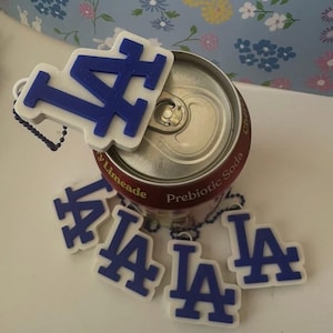 Puede incluir: Llaveros blancos y azules con el logotipo de LA y una lata de refresco. Los llaveros tienen la forma del logotipo de los Dodgers de Los Ángeles. La lata de refresco es roja con texto blanco. El fondo es un estampado floral azul claro.