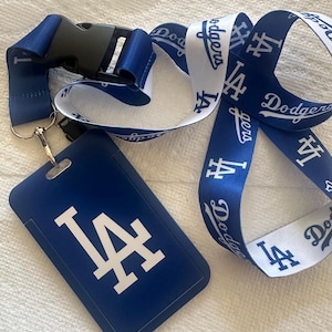 Puede incluir: Un cordón azul y blanco con el logotipo y el texto de los Dodgers de Los Ángeles. El cordón tiene una hebilla desmontable y un soporte para identificación azul con el logotipo del equipo. El cordón es de material sedoso.