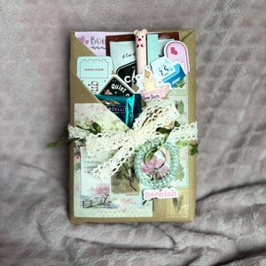 Puede incluir: Una caja de regalo envuelta con encaje y adornos florales. Contiene pegatinas, un bolígrafo y un anillo. La caja lleva la palabra "Bookish". La paleta de colores es suave, con rosas, marrones y blancos.