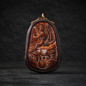 以下が含まれることがあります： 詳細な彫刻が施されたドラゴンデザインの茶色の革製小銭入れ。 ドラゴンはオレンジと茶色の色合いで、白いアクセントが付いています。 小銭入れには、金色の留め金とジッパーが付いています。