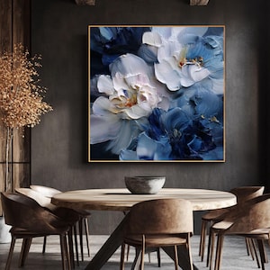 Peut inclure: Peinture florale encadrée avec une bordure dorée. L'œuvre présente de grandes fleurs texturées dans des tons de blanc et de bleu, avec des touches de jaune et d'orange. La peinture est exposée au-dessus d'une table à manger en bois avec des chaises marron.