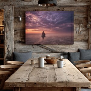 May include: A rustic dining room with a large, weathered wooden table set for a meal. A canvas print of a person on horseback at sunset hangs on the wall. The room features wooden beams and a dark metal pendant light.