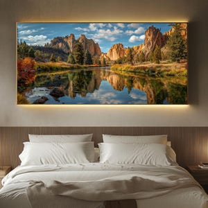 May include: A framed landscape art print featuring a scenic view of a river reflecting the sky and surrounding rock formations. The artwork is displayed above a bed with white linens and a beige throw blanket. The frame has a warm, golden glow.