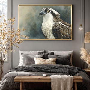 Puede incluir: Una pintura en acuarela enmarcada de un águila pescadora, con una representación detallada de la cabeza y el pecho del ave. La obra de arte está ambientada sobre un fondo tenue y atmosférico con un marco dorado. La pintura se exhibe sobre una cama con ropa de cama gris.