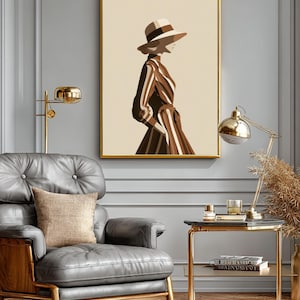 May include: A framed art print featuring a woman in a hat and striped dress, rendered in shades of brown and beige. The artwork is displayed in a gold frame, adding a touch of elegance. The image is set in a room with a gray armchair and gold accents.