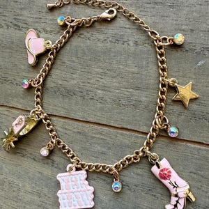 May include: Gold-tone charm bracelet featuring a pink cowboy hat, spur, boot, star, and a charm that says "Yee Haw". The bracelet has iridescent crystal accents and a lobster clasp closure. The charms are detailed with pink and gold enamel.