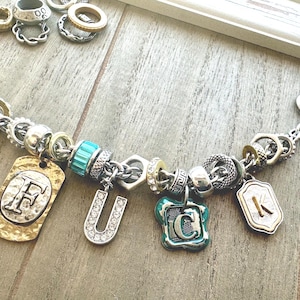 May include: A silver charm bracelet with various charms, including letter pendants and decorative beads. The bracelet features a toggle clasp and a mix of silver, gold, and turquoise accents. The charms include the letters F, U, C, and K.