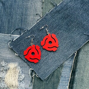 May include: A pair of red acrylic earrings in the shape of 45 RPM record adapters. The earrings hang from silver-colored fish hook ear wires. The earrings are displayed on a background of blue denim fabric.