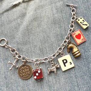 May include: Silver-toned charm bracelet featuring various game-themed charms. Charms include a domino, playing card, chess piece, Scrabble tile, dice, dog, cross, and a coin with "NO CASH VALUE" text. The bracelet is displayed on a denim background.