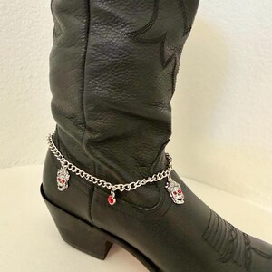 May include: A black leather cowboy boot with a silver chain ankle bracelet. The bracelet features skull charms with red accents and a small red gem. The boot has decorative stitching.