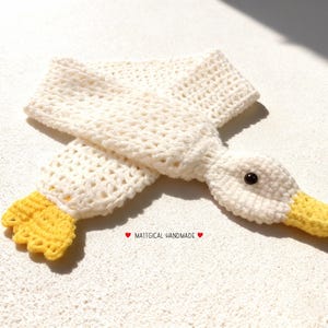 May include: A handmade, off-white crochet scarf shaped like a goose. The scarf features a goose head with a yellow beak and black eye, and yellow feet. The text "MATTICAL HANDMADE" is also visible.