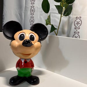 Puede incluir: Una figura de Mickey Mouse con cabeza oscilante vintage. La figura tiene una cara bronceada, orejas negras y una camisa roja con pantalones verdes. La figura está sobre una superficie blanca.