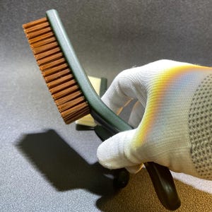 K&ouml;nnte beinhalten: Eine gr&uuml;n-griffige B&uuml;rste mit braunen Borsten. Die B&uuml;rste wird in einer wei&szlig; behandschuhten Hand gehalten. Die Borsten sind dicht gepackt und scheinen aus synthetischem Material zu bestehen. Der Griff hat ein gebogenes Design f&uuml;r einen einfachen Griff.
