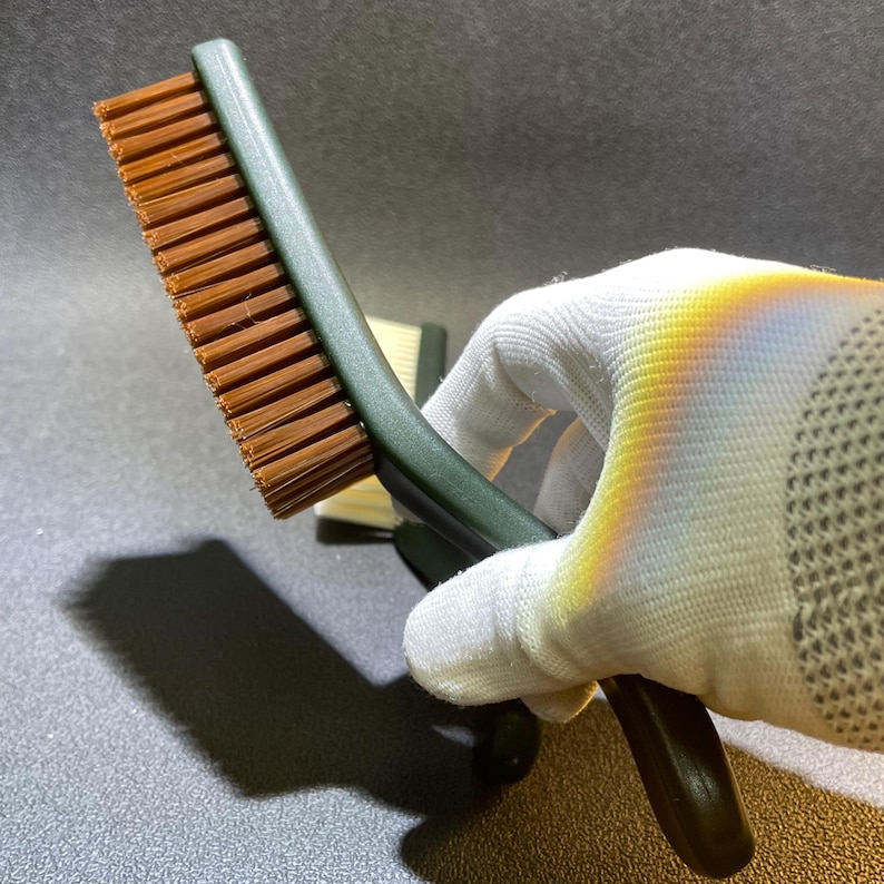 K&ouml;nnte beinhalten: Eine gr&uuml;n-griffige B&uuml;rste mit braunen Borsten. Die B&uuml;rste wird in einer wei&szlig; behandschuhten Hand gehalten. Die Borsten sind dicht gepackt und scheinen aus synthetischem Material zu bestehen. Der Griff hat ein gebogenes Design f&uuml;r einen einfachen Griff.
