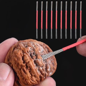May include: Close-up of a brown walnut being worked on with a tool. The tool has a red handle and a checkered tip. Several similar tools are arranged vertically in the background against a black backdrop.