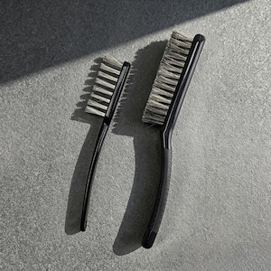 May include: Two black handled brushes with silver bristles are displayed on a gray surface. One brush is smaller with a straight handle, while the other is larger with a curved handle. The bristles are tightly packed.