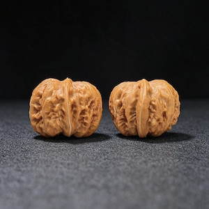 May include: Two light brown walnuts with deeply textured, wrinkled shells. Each nut features a prominent, raised central ridge running vertically along the surface, showcasing the natural, intricate patterns and organic contours of the hard outer casing.
