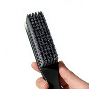 May include: A black rectangular brush with closely packed, dark gray bristles. The brush has a black handle and a light green edge. The bristles are densely packed, creating a textured surface. The brush is held in a hand.