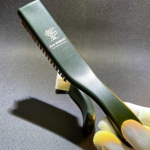 K&ouml;nnte beinhalten: Eine dunkelgr&uuml;ne B&uuml;rste mit goldenen Borsten und einem gebogenen Griff. Auf der B&uuml;rste steht "Zen Walnut The Patina Project" und ein Baumlogo auf dem Griff.