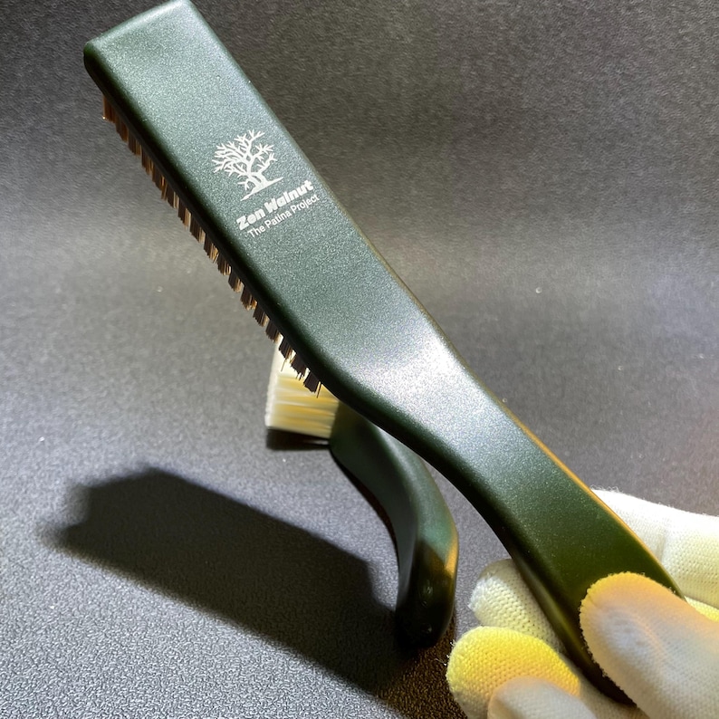 K&ouml;nnte beinhalten: Eine dunkelgr&uuml;ne B&uuml;rste mit goldenen Borsten und einem gebogenen Griff. Auf der B&uuml;rste steht "Zen Walnut The Patina Project" und ein Baumlogo auf dem Griff.