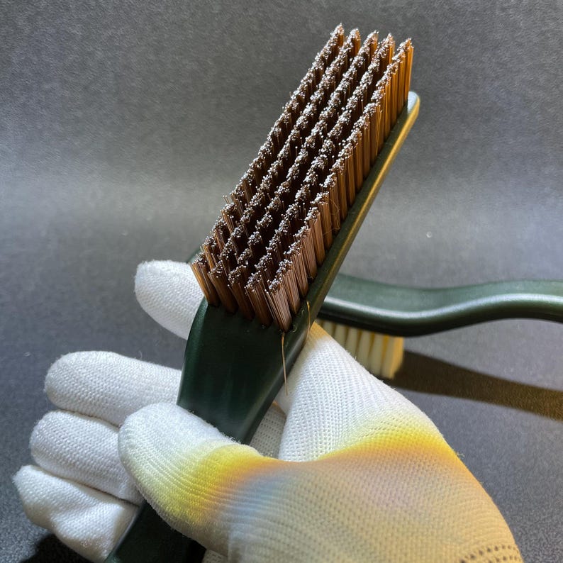 K&ouml;nnte beinhalten: Nahaufnahme einer dunkelgr&uuml;nen B&uuml;rste mit braunen Borsten. Die B&uuml;rste wird von einer behandschuhten Hand gehalten. Die Borsten sind in einem rechteckigen Muster angeordnet. Eine weitere B&uuml;rste ist im Hintergrund sichtbar.