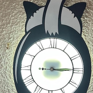 Puede incluir: Un reloj de pared caprichoso con forma de gato, con una esfera blanca con números romanos y manecillas negras. El cuerpo del gato es azul oscuro con detalles blancos. El reloj está montado en una pared texturizada.