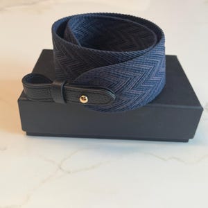 May include: A coiled navy blue camera strap featuring a dark chevron pattern, finished with black textured leather end tabs and a single gold-toned metal rivet, resting on top of a rectangular black box.