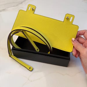 May include: A bright yellow leather phone case with a matching strap, open to reveal a black interior. The case has a rectangular shape with rounded corners and gold-colored hardware. The strap is thin and appears to be adjustable.