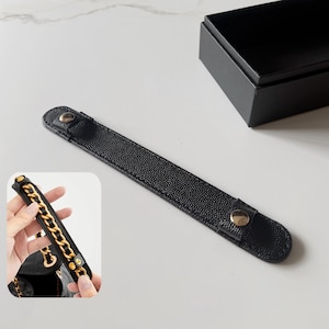 May include: A black textured leather strap with gold-tone metal snap buttons at each end. The strap is designed to wrap around a handbag chain, as shown in the inset image, providing a comfortable grip with its pebbled surface and secure fasteners.