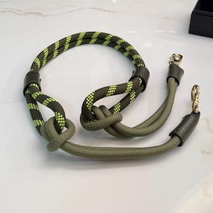 May include: A dog leash and collar set. The collar is olive green with neon yellow accents, knotted at the center. The leash is a matching olive green with gold-colored metal clasps and leather accents. The set is designed for pet owners.