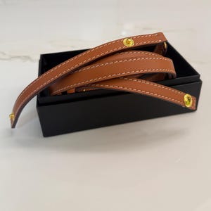 Puede incluir: Una correa de cuero marrón claro con costuras blancas y herrajes dorados está enrollada dentro de una caja negra. La correa parece ser un asa o un accesorio para un bolso u otro artículo. El cuero tiene un acabado liso.