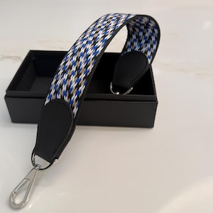 May include: A patterned bag strap with a houndstooth design in blue, white, and tan. The strap has black leather ends and silver-tone metal clasps. The strap is displayed on a white surface with a black box in the background.