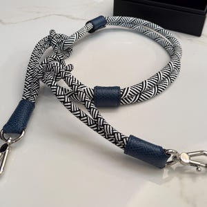 May include: A black and white patterned rope strap with navy blue leather accents and silver-tone metal clasps. The strap is designed with a woven pattern and is suitable for a bag or other accessories.