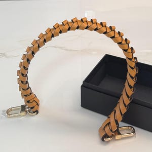 May include: A tan leather braided purse handle with gold-colored clasps. The handle is arched and sits next to a black box on a white surface. The leather is a warm, light brown color.